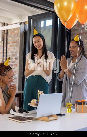 Diverse colleghe che applaudono le mani per festeggiare il compleanno intorno a un tavolo bianco tramite laptop Foto Stock