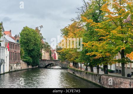 Un pittoresco canale di Bruges, Belgio, caratterizzato da un vecchio ponte in pietra e alberi autunnali con foglie d'arancio che si riflettono nell'acqua, catturando il fascino Foto Stock