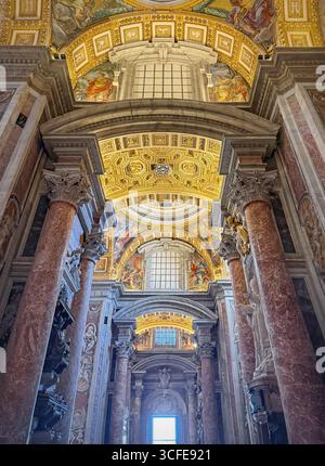 Una vista che guarda all'interno della maestosa Basilica di San Pietro. Enormi colonne in marmo rossastro incorniciano il soffitto ornato, riccamente decorato con patte dorate Foto Stock