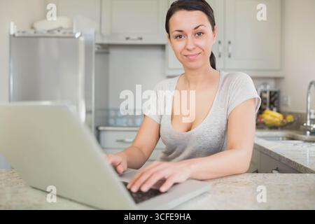 Donna che scrive su un computer portatile argentato in un'isola di cucina in pietra con lavandino cromato, banane e frigorifero Foto Stock