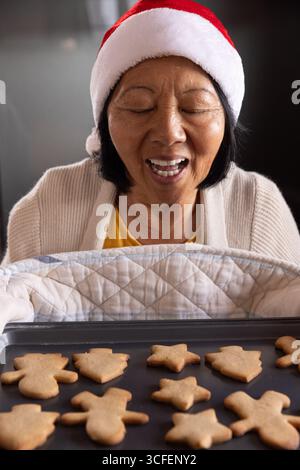 Donna asiatica anziana che indossa il cappello di Babbo Natale tenendo il vassoio al banco della cucina che soffia sui biscotti delle vacanze Foto Stock