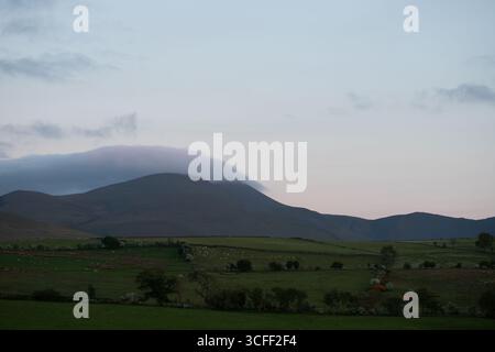 Un tranquillo paesaggio crepuscolare caratterizzato da dolci montagne parzialmente avvolte da morbide nuvole sospese. Sotto, ampi campi verdi si estendono verso l'esterno Foto Stock