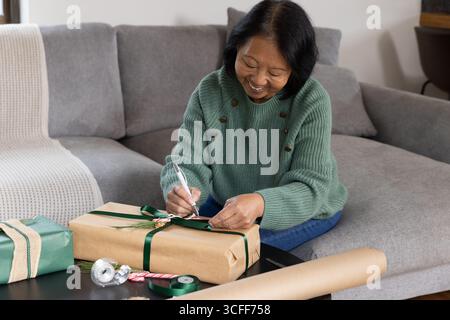 Donna asiatica anziana che confeziona il regalo sul tavolino da caffè a casa usando forbici, nastro e nastro Foto Stock