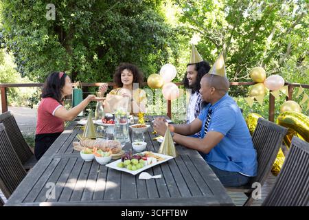 Diversi amici seduti intorno al tavolo sul ponte condividendo una borsa regalo, assaggiando dolci in mezzo a palloncini Foto Stock