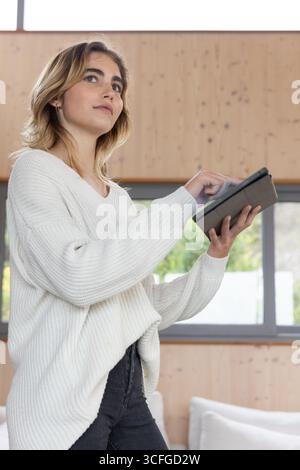Donna che utilizza un tablet nero in un salotto moderno con pareti rivestite in legno e divano imbottito bianco Foto Stock