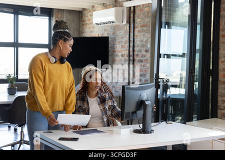 Collaboratrici afroamericane che collaborano alla progettazione alla scrivania dell'ufficio con monitor e documenti Foto Stock