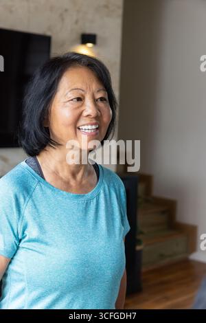 Donna asiatica anziana in piedi in un moderno salotto con top atletico e sorridente vicino alla televisione Foto Stock