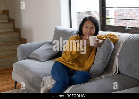 Donna asiatica anziana seduta sul divano grigio in soggiorno tenendo la tazza e appoggiandosi sulla coperta Foto Stock