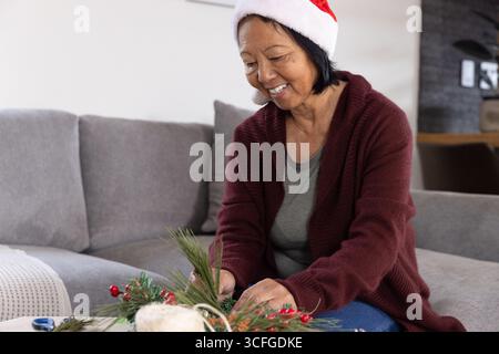 Donna asiatica anziana che indossa il cappello di babbo natale che organizza rami di pino e frutti di bosco in un pezzo centrale Foto Stock