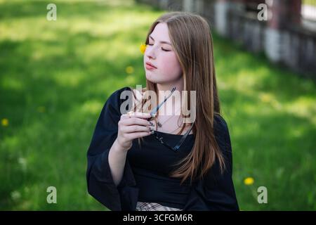 Studentessa delle scuole superiori che si rilassa in un parco durante le vacanze primaverili, tenendo gli occhiali da sole e con un dente di leone sulla guancia Foto Stock