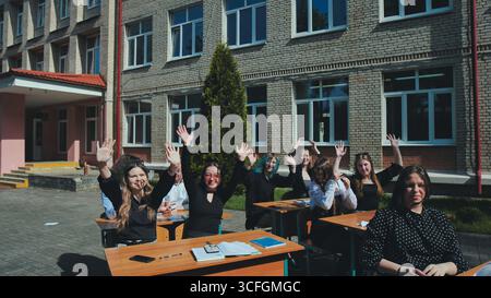 Studenti felici che alzano le mani alle scrivanie all'aperto, si divertono molto durante la giornata scolastica Foto Stock