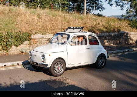 Fiat 500 d'epoca bianca classica 1971 guidata da un uomo su una strada soleggiata con pareti in pietra e vegetazione lussureggiante sullo sfondo Foto Stock
