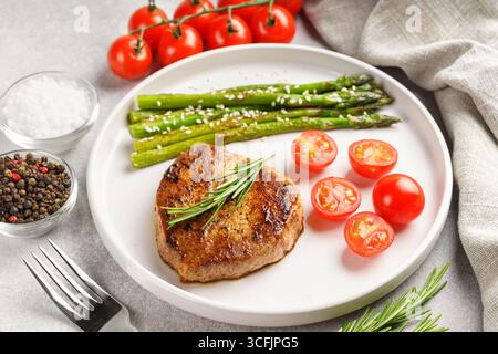 Primo piano, macina di gustosa bistecca di manzo alla griglia servita con asparagi verdi e pomodori ciliegini su piatto bianco, con posate. Perfetto per menu, dieta, Foto Stock