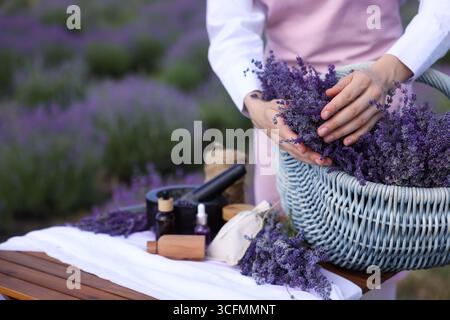 Fare cosmetici naturali. Donna con cesto in vimini di fiori di lavanda e altri ingredienti a tavola nel campo, primo piano. Spazio per il testo Foto Stock