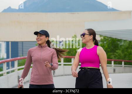 Due giovani donne latine in abbigliamento sportivo sorridono e camminano insieme su un percorso all'aperto, godendo di un'attività piacevole Foto Stock