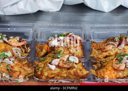 Omelette di ostriche thailandesi con gamberi e calamari, marrone dorato e servita in un contenitore di plastica trasparente, un popolare piatto di Street food thailandese. Foto Stock