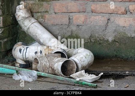 Una pila di rottami di vecchio tubo in PVC rotti e un tubo dell'acqua danneggiato si trovano su un tetto di cemento sporco e bagnato coperto di muschio, con una scatola di schiuma scartata nelle vicinanze. Foto Stock
