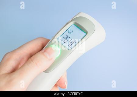 Mano femmina che regge un moderno termometro a infrarossi isolato su sfondo blu. Misurazione remota della temperatura corporea. Controllo delle malattie infettive Foto Stock