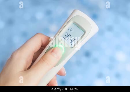 Mano femminile che tiene in mano un moderno termometro a infrarossi isolato su fondo in tessuto blu. Misurazione a distanza della temperatura corporea. Controllo dell'infettio Foto Stock