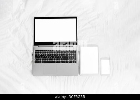 laptop, tablet e telefono con schermo bianco su un letto bianco. Notebook grigio aperto con tablet digitale bianco e smartphone su sfondo bianco. finta Foto Stock