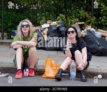 Due spettatori femminili siedono sul marciapiede con i rifiuti dietro durante il Carnevale di Notting Hill, Londra. Inghilterra 25 agosto 2025 Foto Stock
