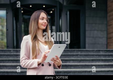 analista finanziaria caucasica che indossa una tuta da lavoro che regge il tablet mentre si trova sullo sfondo di un edificio per uffici in città Foto Stock