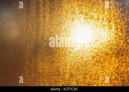 Consistenza dorata e astratta di goccioline d'acqua condensata sulla superficie della finestra illuminata da dietro dalla luce del sole. Foto Stock