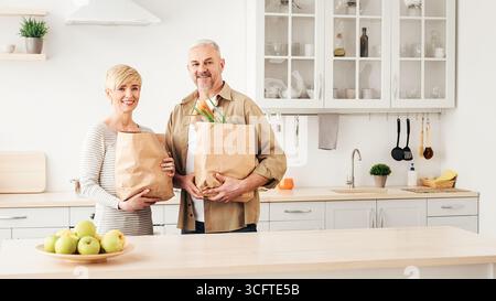 Felice coppia sposata che viene a casa dallo shopping e dalla disimballaggio della carta borse con generi alimentari in cucina insieme Foto Stock