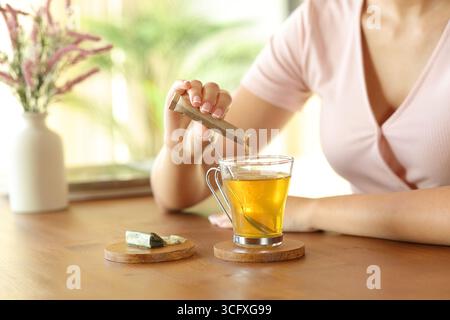 Donna che versa a mano zucchero nel bicchiere da tè su un tavolo di legno a casa Foto Stock