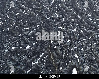 Vista aerea di un paesaggio aspro e aspro dove la terra vulcanica scura incontra macchie di neve sparse, creando un mosaico testurizzato, le Highlands, Islanda Foto Stock