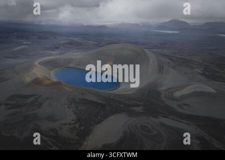 Veduta aerea di un suggestivo lago cratere blu annidato tra gli aspri altopiani vulcanici, in contrasto con il terreno oscuro e accidentato, gli altipiani e l'Islanda. Foto Stock