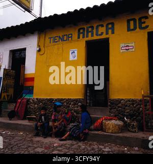 Zwei Frauen und ein Junge sitzen vor einem gelb gestrichenen Laden mit der Aufschrift „Tienda Arca de…“, neben Körben und Waren. Guatemala, um 1980. Foto Stock