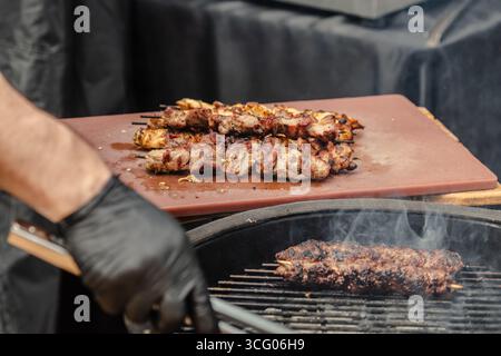 Uno shot ravvicinato di una persona che grigia spiedini di carne su un barbecue, indossa guanti neri, usa pinze per capovolgere la carne mentre il fumo fuoriesce dalla griglia Foto Stock