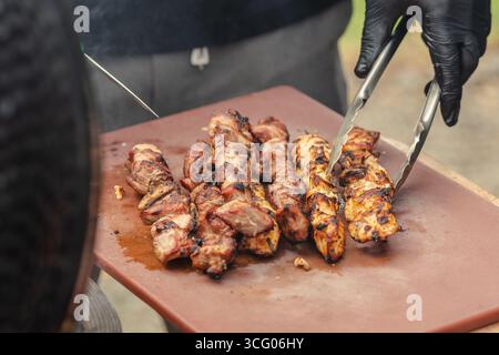 Uno shot ravvicinato di una persona che grigia spiedini di carne su un barbecue, indossa guanti neri, usa pinze per capovolgere la carne Foto Stock