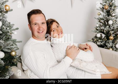 Coppia felice in maglioni bianchi seduti insieme in un accogliente studio natalizio con alberi decorati e atmosfera festosa. Foto Stock