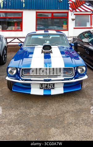 Una classica muscle car Ford Mustang a righe bianche e blu degli anni '1960 è parcheggiata di fronte a una tavola calda, che mostra il suo design vintage e il suo potente motore. Foto Stock