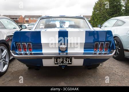 Vista posteriore di un'auto Ford Mustang vintage a righe bianche e blu parcheggiata tra due veicoli moderni in una giornata nuvolosa Foto Stock