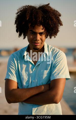 Giovane con i capelli afro sorridente con le braccia piegate sulla spiaggia Foto Stock