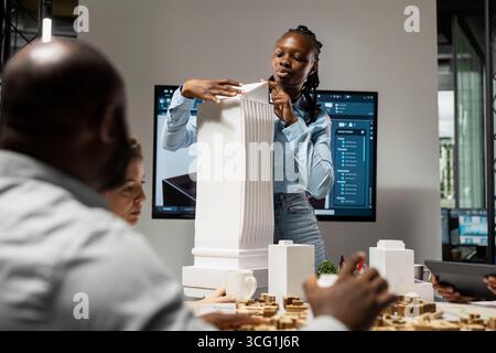 L'appaltatore afroamericano mostra modelli 3D architettonici per il futuro cantiere, lavoro di squadra per un nuovo progetto immobiliare. Piano generale per la progettazione di un progetto residenziale, pianificazione urbana. Foto Stock