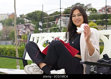 Giovane studente universitario indiano seduto su una panchina all'aperto, leggendo un libro e mangiando un croissant, simboleggiando lo stile di vita accademico moderno, l'equilibrio di stu Foto Stock