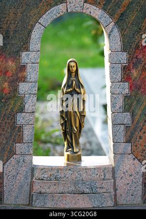 La piccola statua dorata della vergine maria con rosario sta pregando in una nicchia di granito di un cimitero Foto Stock