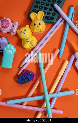 Penne color pastello, matite, gomme, affilatrici e una calcolatrice creano un'allegra scena di ritorno a scuola su un vivace sfondo arancione Foto Stock