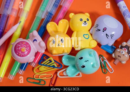 Graziosi strumenti di affilatura a forma di animale, penne colorate e graffette creano una disposizione vivace su uno sfondo arancione Foto Stock