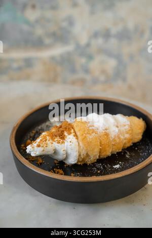 Primo piano di un gustoso dessert di pasta sfoglia ripieno di panna montata dolce spolverata di briciole di caramello, in un piatto di ceramica nera. Foto Stock