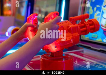 Primo piano di una mano che regge una pistola giocattolo rossa dall'aspetto futuristico di un gioco arcade. L'emozione e l'azione di giocare a un videogame in una sala giochi Foto Stock