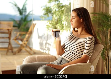 Donna a casa seduta su una poltrona che beve un caffè guardando lontano da una finestra di casa Foto Stock
