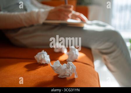 Una donna siede su un divano arancione, scrive su un notebook e scarta pezzi di carta sgualciti, suggerendo difficoltà creative o processo di editing Foto Stock