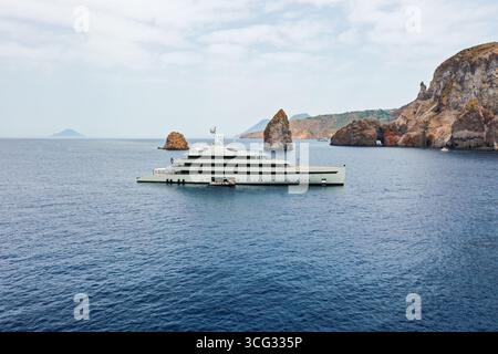 Vista aerea di un elegante e moderno yacht che naviga sulle acque blu profonde vicino alle aspre coste rocciose delle Isole Eolie, Sicilia, Italia. Foto Stock