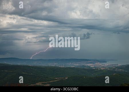 Un fulmine maestoso colpisce il cielo nuvoloso sul paesaggio urbano costiero tra Trieste, Italia e Capodistria, Slovenia. Foto Stock