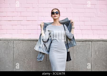 Moderna donna urbana che indossa una giacca in denim e occhiali da sole su una parete rosa pastello. Ritratto di Street fashion con eleganza informale e alla moda. Perfetto Foto Stock